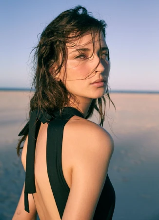 Close-up portrait of a woman with wet dark hair in a black halter top against a sandy beach and clear sky.