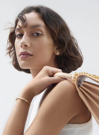 Close up portrait of a woman with short dark wavy hair holding a pale pink shoulder bag with a gold chain