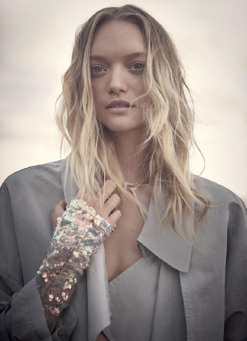 Portrait of a blonde female model in a grey coat and sequined glove, standing outdoors with a blurred light grey background.