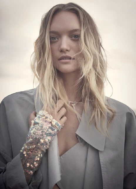 Portrait of a blonde female model in a grey coat and sequined glove, standing outdoors with a blurred light grey background.