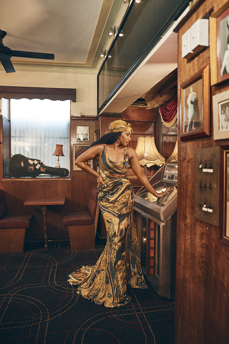 Elegant adult woman in patterned gown poses indoors near jukebox, accessorized with a matching headscarf and earrings.