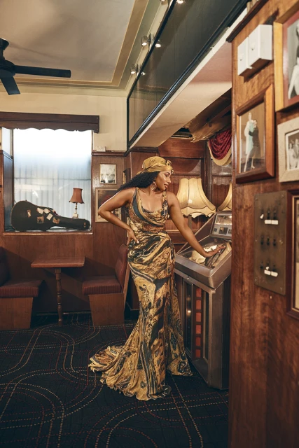 Elegant adult woman in patterned gown poses indoors near jukebox, accessorized with a matching headscarf and earrings.