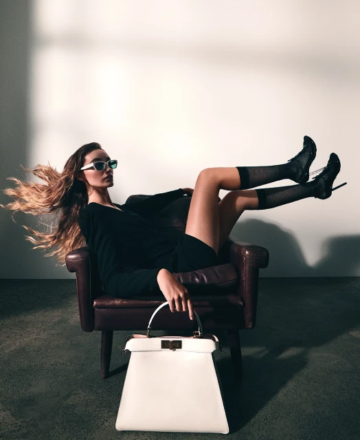 A fashionable woman poses in an armchair with legs up wearing sunglasses, a dress, heels, and holding a white purse in a studio.