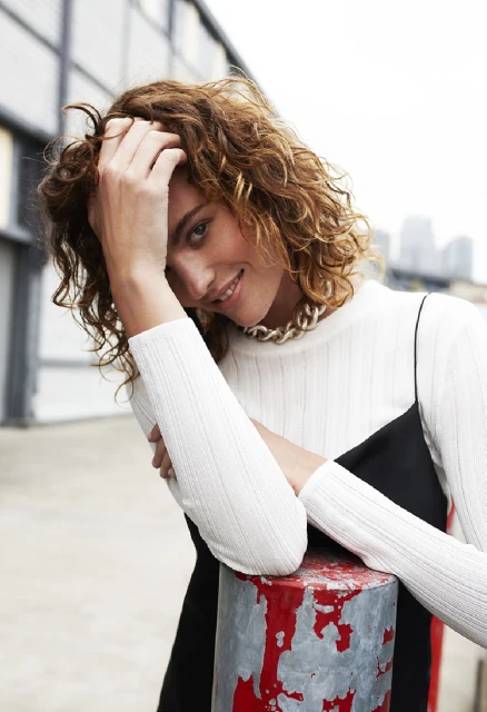 Portrait of a smiling woman with curly brown hair wearing a white long-sleeved shirt and a black dress outdoors with blurred background.
