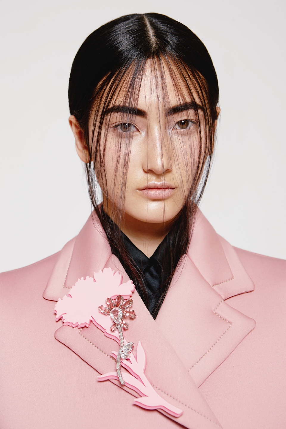 Headshot of a woman with black hair wearing a light pink jacket, featuring a pink flower brooch, against a white backdrop.