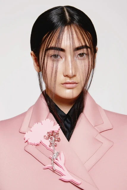 Headshot of a woman with black hair wearing a light pink jacket, featuring a pink flower brooch, against a white backdrop.