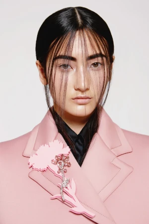 Headshot of a woman with black hair wearing a light pink jacket, featuring a pink flower brooch, against a white backdrop.