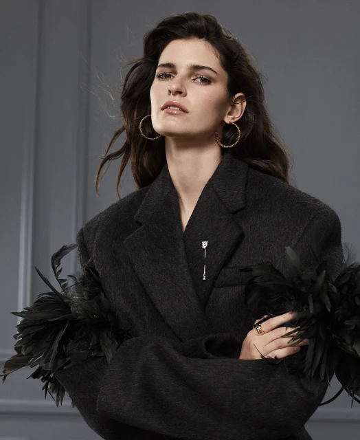 Portrait of a brunette woman wearing a dark blazer with feather details, hoop earrings, and ring, posing against a grey background.