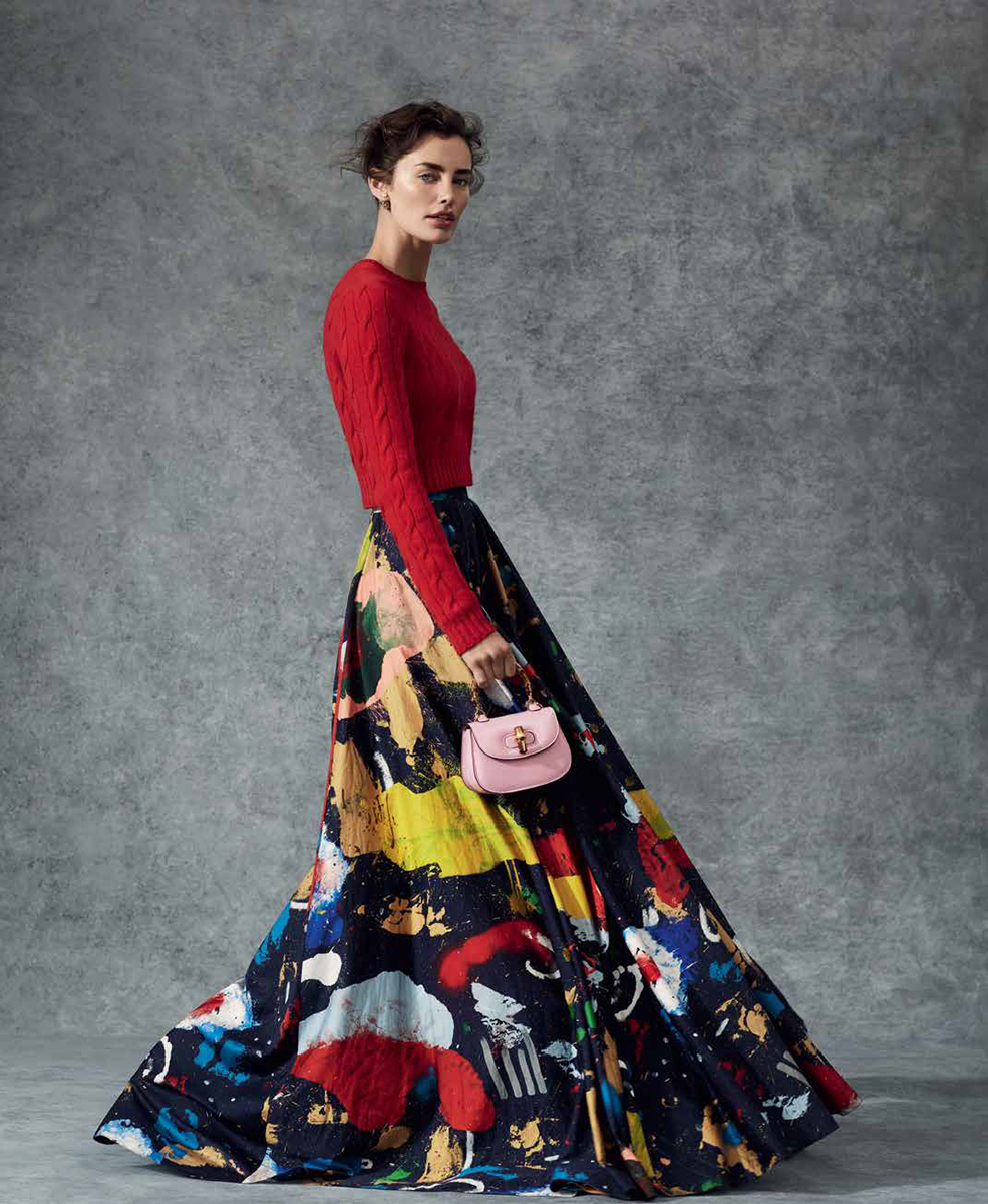 Fashionable woman poses in a vibrant maxi skirt and red sweater, complemented by a petite pink handbag against a textured grey backdrop.