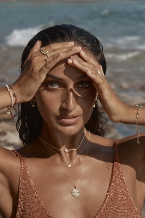 Close-up portrait of a woman wearing gold layered necklaces and rings, with wet hair against an ocean background