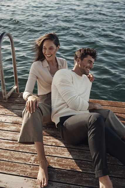 Smiling couple sits on a wooden pier by the sea on a sunny day. The woman has dark hair and wears a beige blouse and pants. The man wears a white sweater.