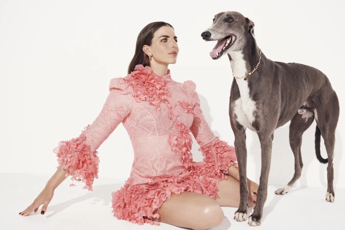 An adult woman in a pink dress poses seated with a greyhound dog in a bright, minimalist photo studio with a white background.