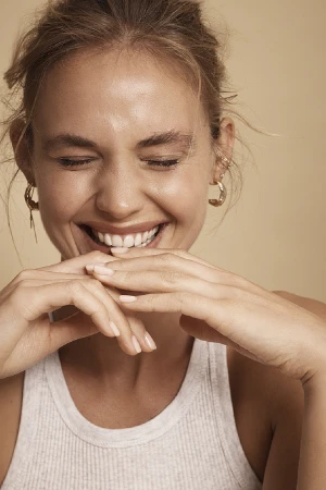 Close-up of a smiling woman with fair skin and blonde hair in a white ribbed tank top. She has hoop earrings and a natural expression.