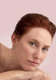 Close up beauty portrait of a red haired woman with light freckles resting her chin on her hands against a pale pink background