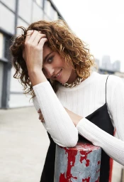 Woman with curly brown hair wearing a white ribbed top and black slip dress leaning against a red pole outdoors.