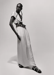 Black model in a long light dress with a wide belt and dark sandals standing against a white seamless backdrop