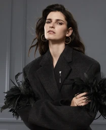 Portrait of a woman with dark hair wearing a black jacket with feathered sleeves and gold hoop earrings