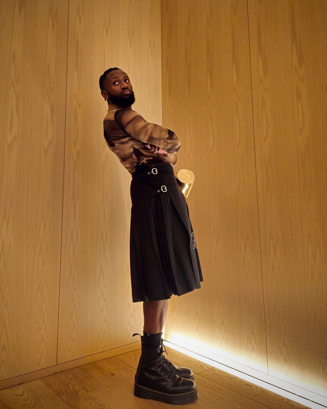 Adult male stands in a black kilt and black boots against a light wood wall in an indoor environment with soft overhead lighting.