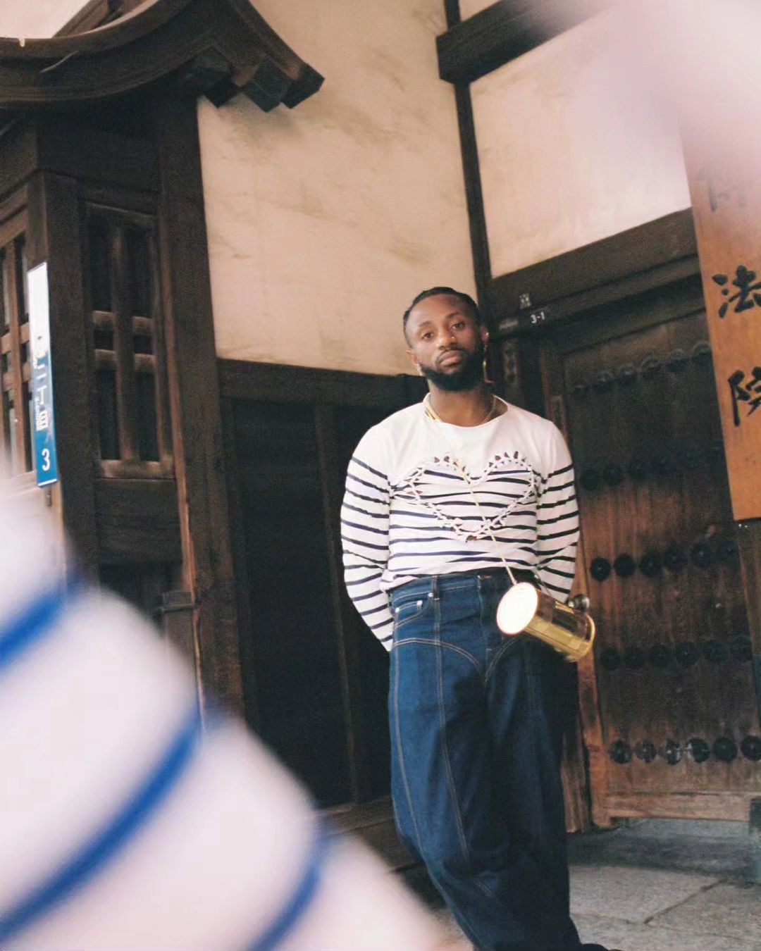 Adult man poses in front of a traditional Japanese building wearing a striped shirt, jeans, and carrying a brass-colored container.
