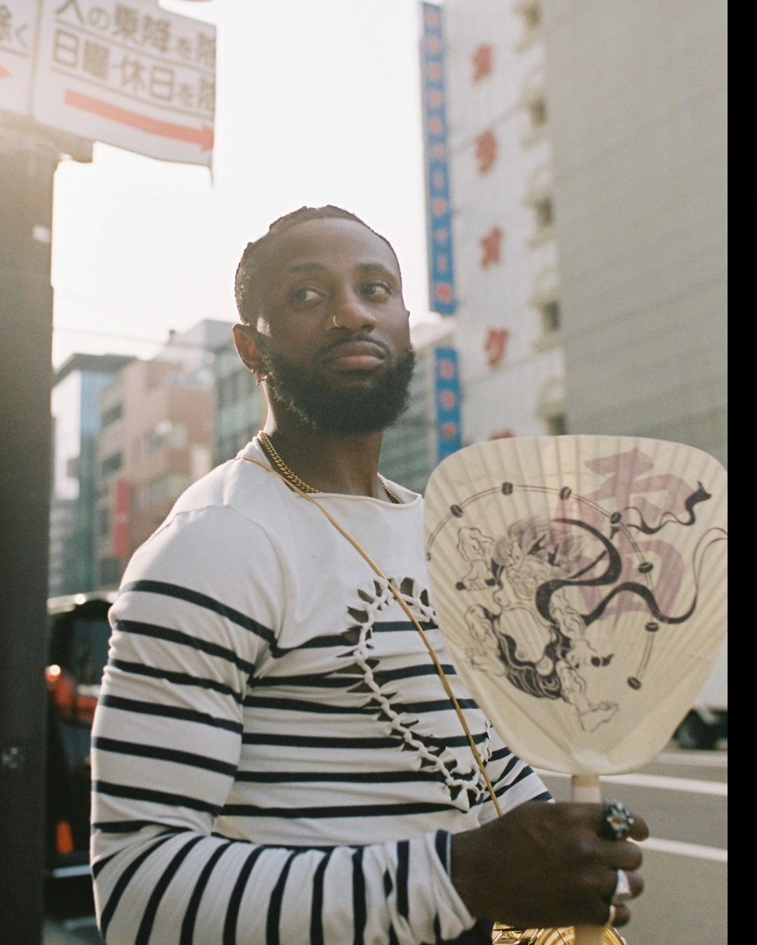 Stylish man in striped shirt and gold chain poses in an urban setting holding a handheld fan with city buildings in the background.