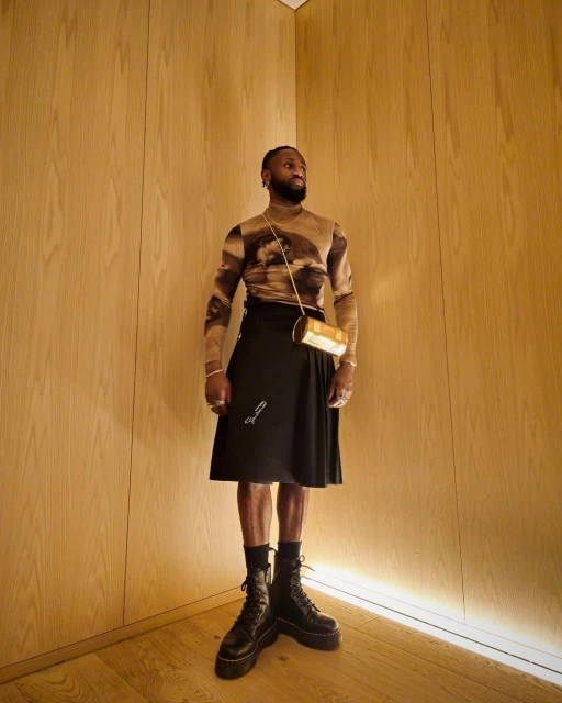 A stylish adult male poses indoors wearing a patterned long-sleeve shirt, black kilt, platform boots, and a gold crossbody bag.
