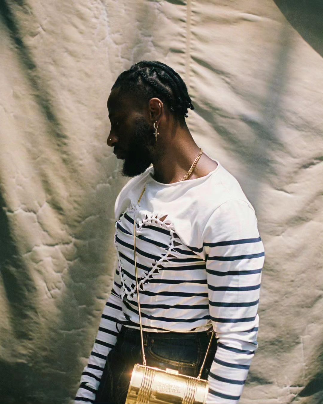 Black male model poses with a striped shirt and gold purse against a textured beige background in a studio portrait.