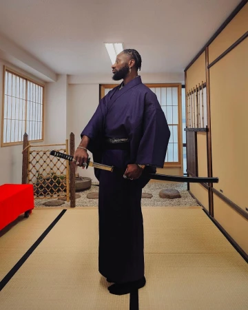 Adult male in traditional Japanese clothing poses indoors holding a katana sword in a room with tatami mats and paper windows.