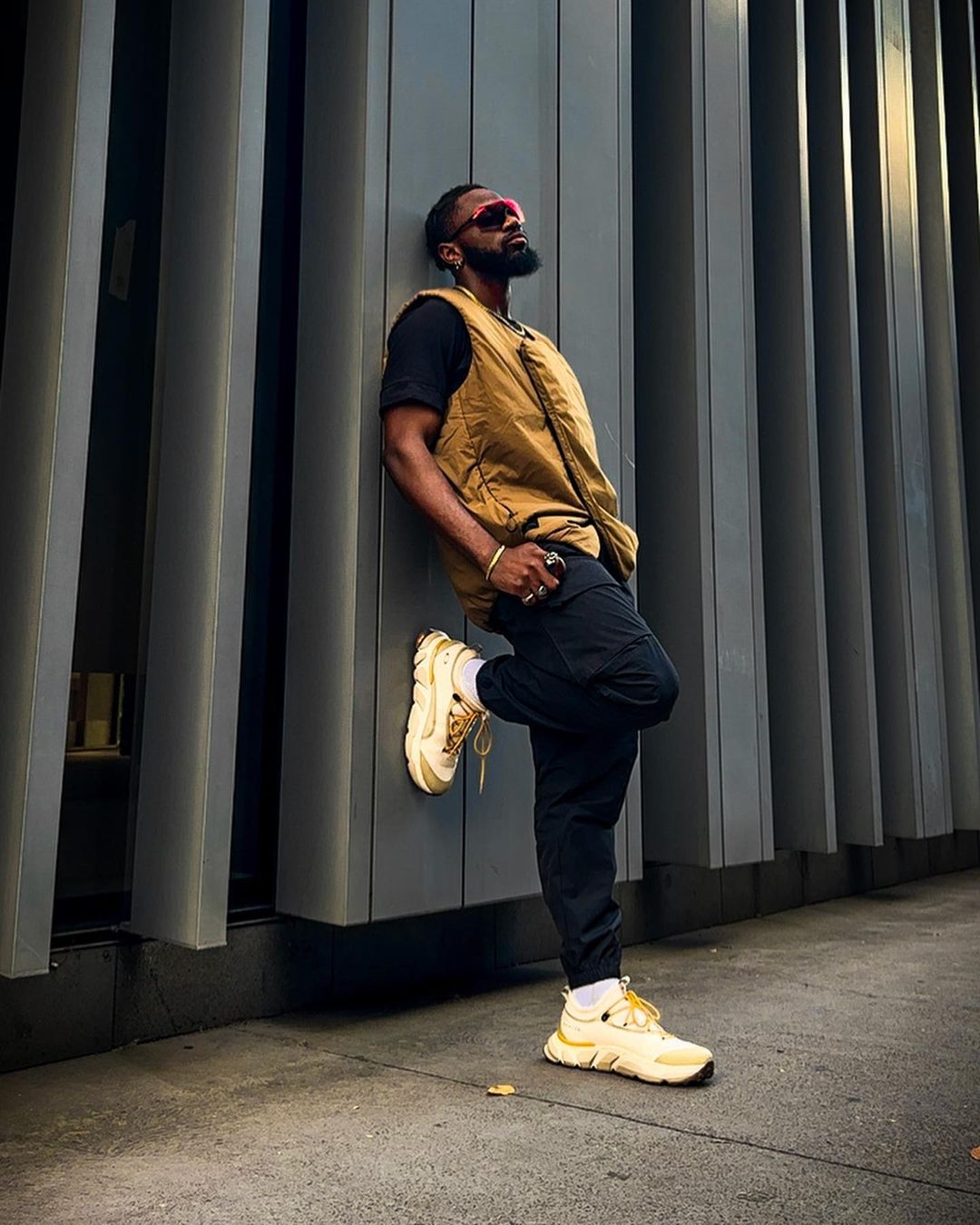 Stylish bearded man with pink glasses poses against modern grey wall wearing a brown vest, black shirt, dark pants and trendy beige sneakers.