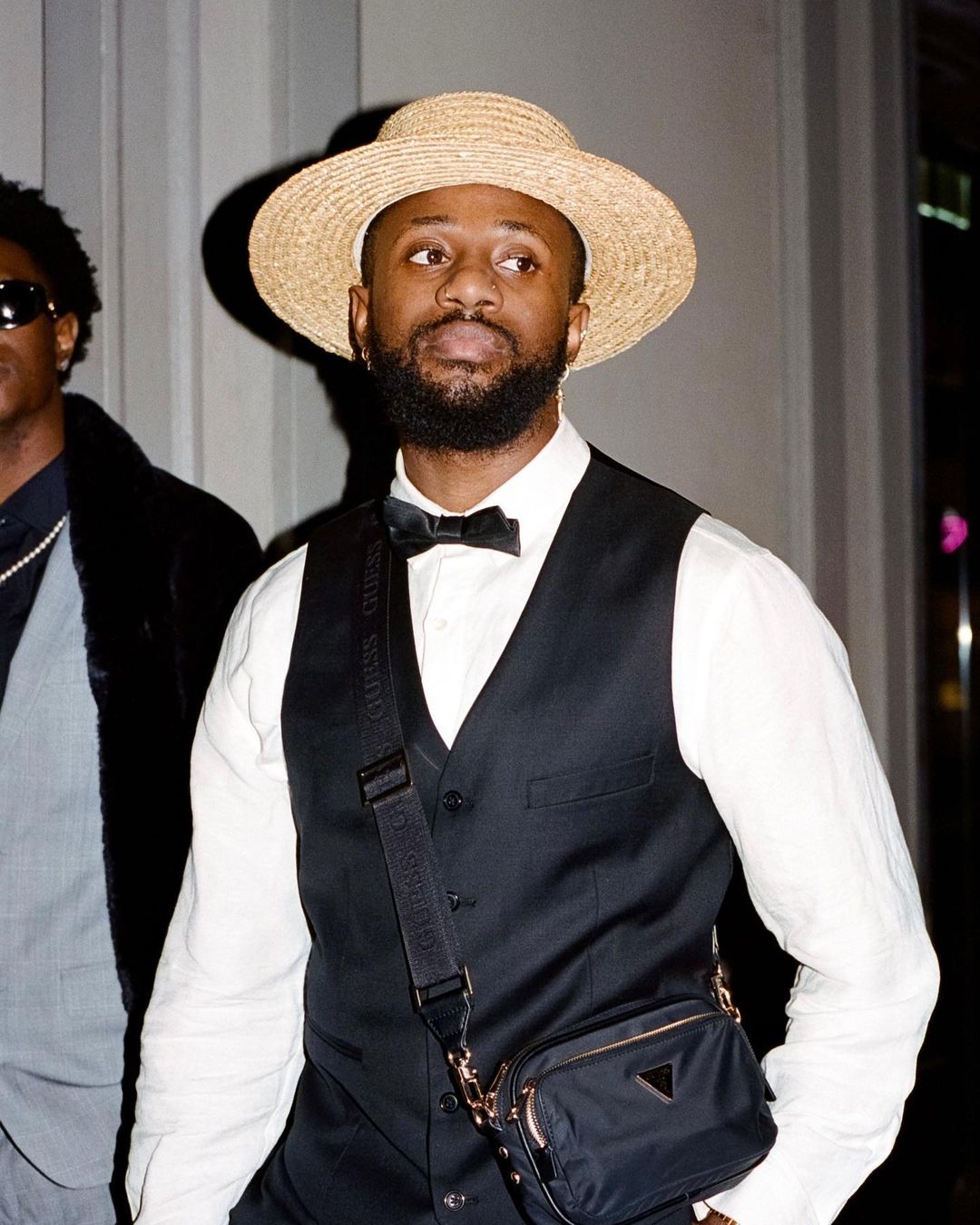 Stylish man in vest, white shirt, bow tie, and straw hat, poses indoors with another man in the background, creating a fashion-forward image.