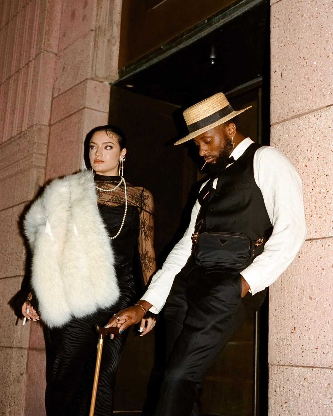 A man and woman in vintage attire pose outside a building at dusk. The woman holds a cane and wears a fur stole.