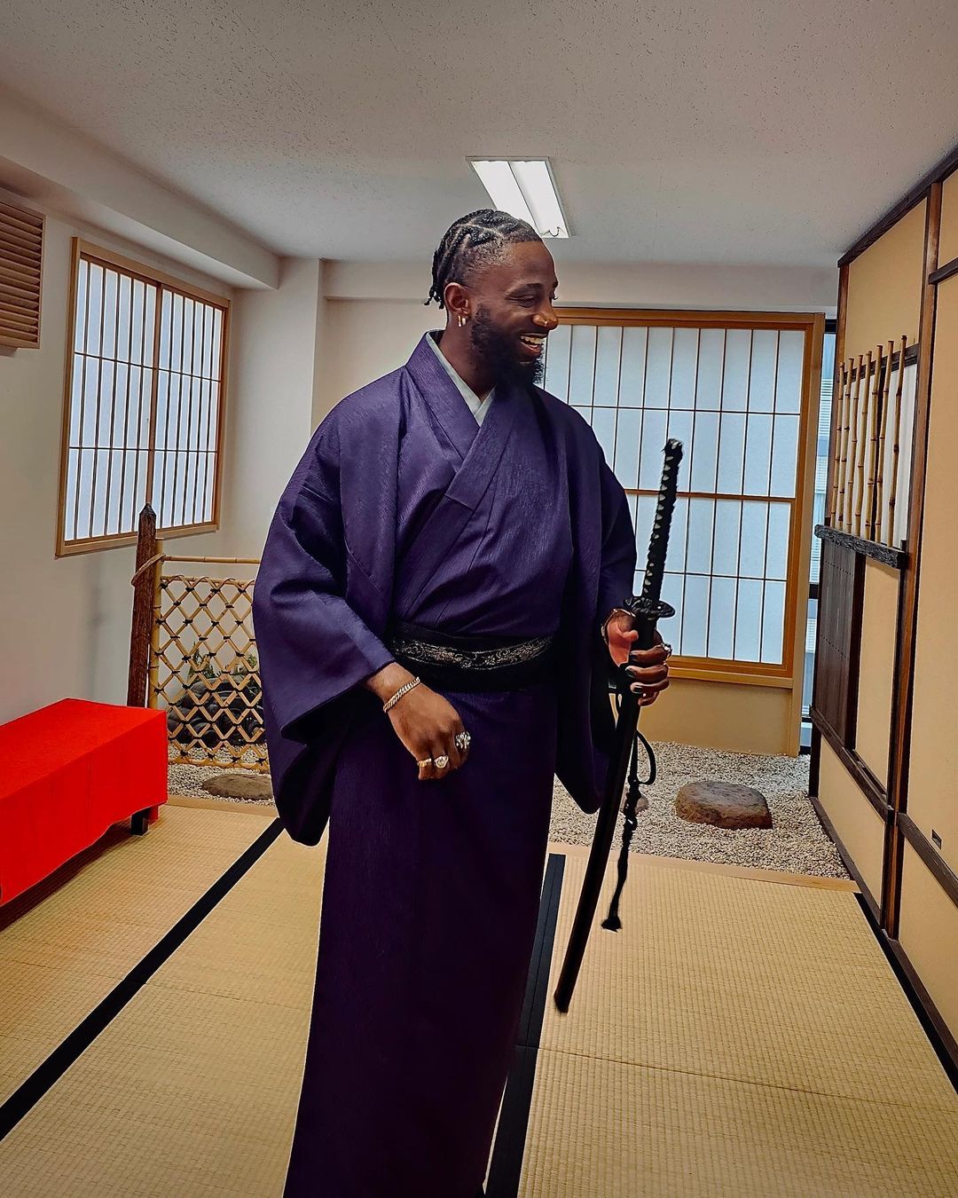 Adult male in purple kimono holding a sword inside an indoor tatami mat room with windows, creating a cultural portrait.