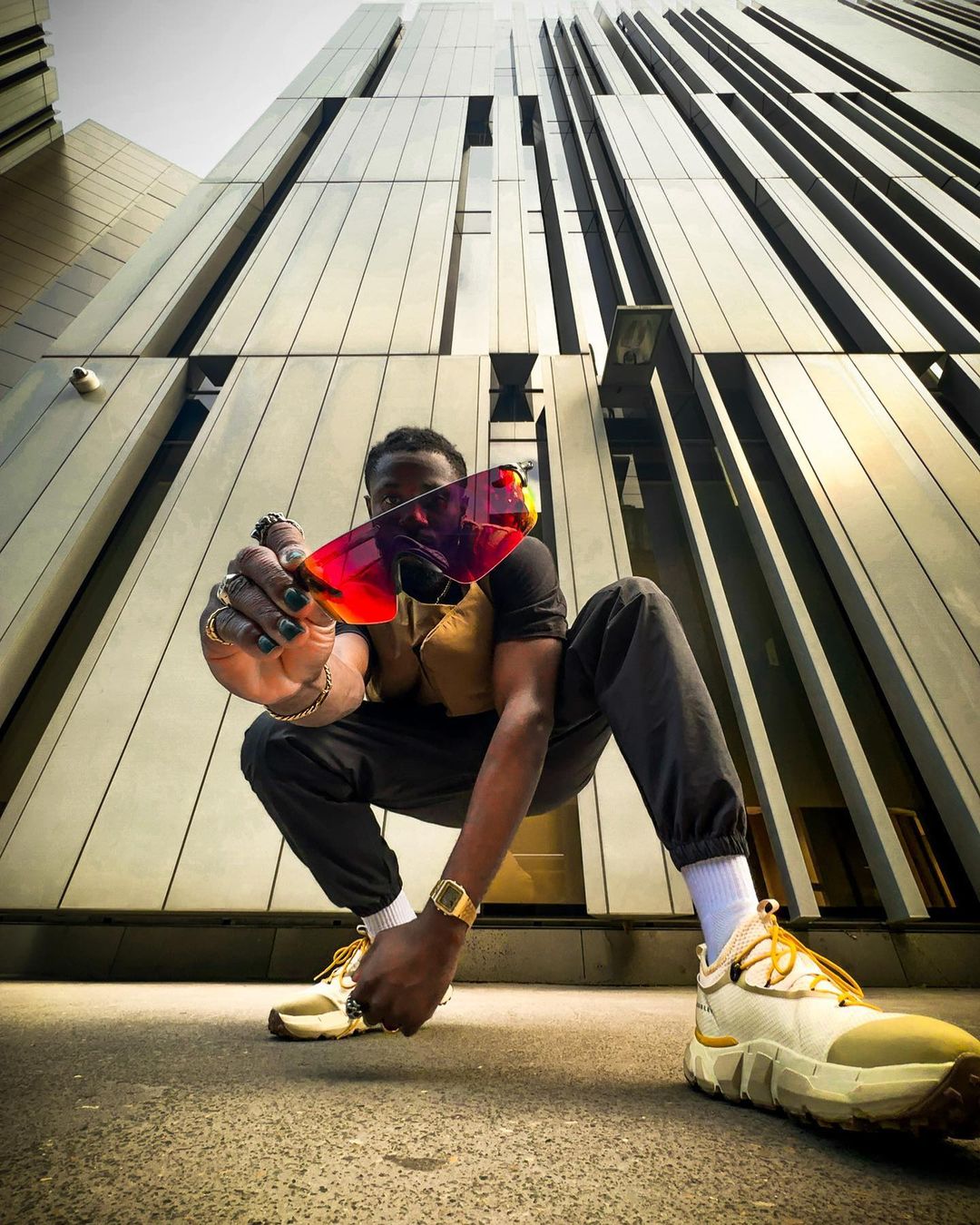 Stylish man in black pants, tee, and sneakers squats while holding red and purple sunglasses. Modern architecture in background.