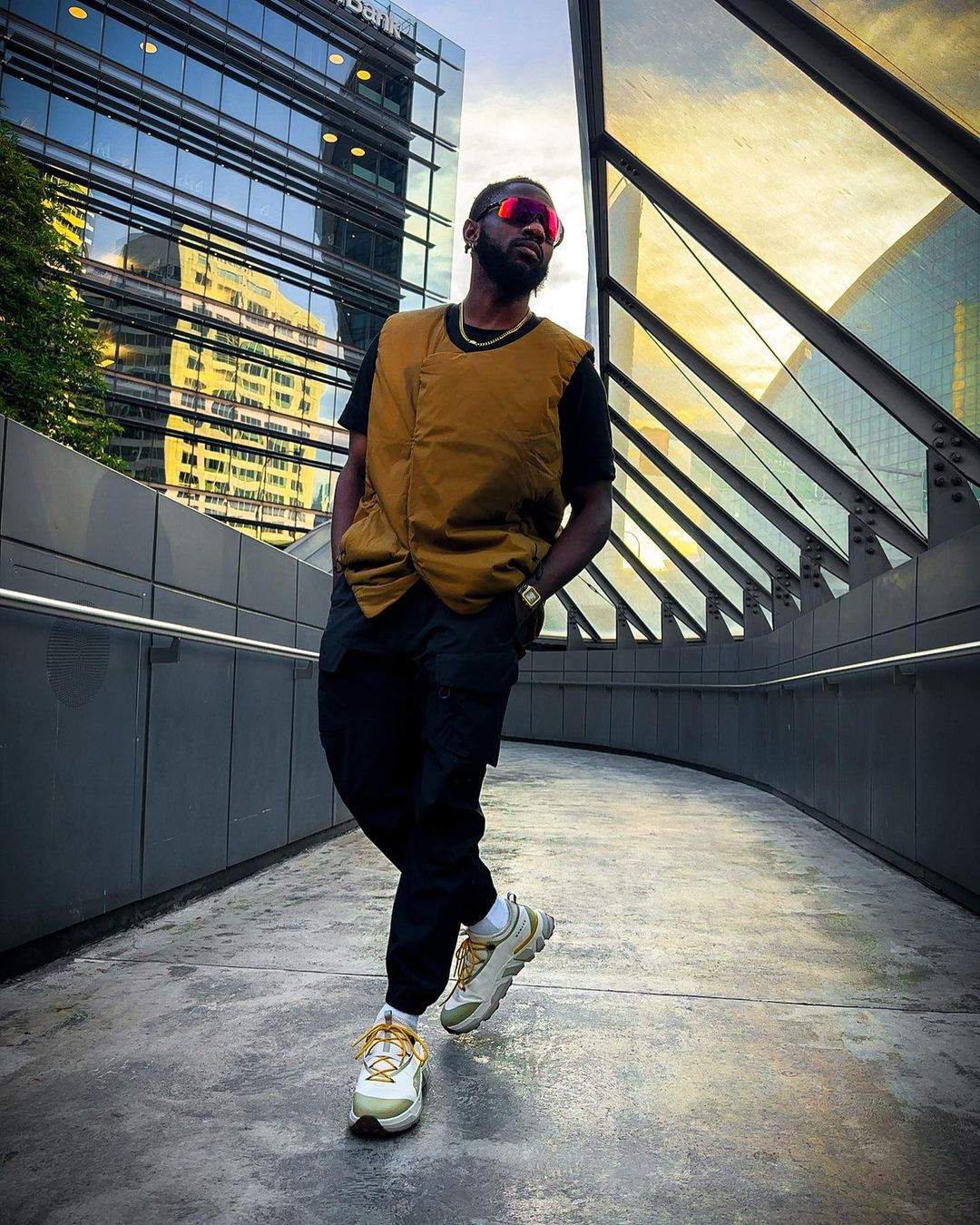Stylish man poses in black pants and vest near modern architecture with red sunglasses and sneakers on an outdoor city walkway.