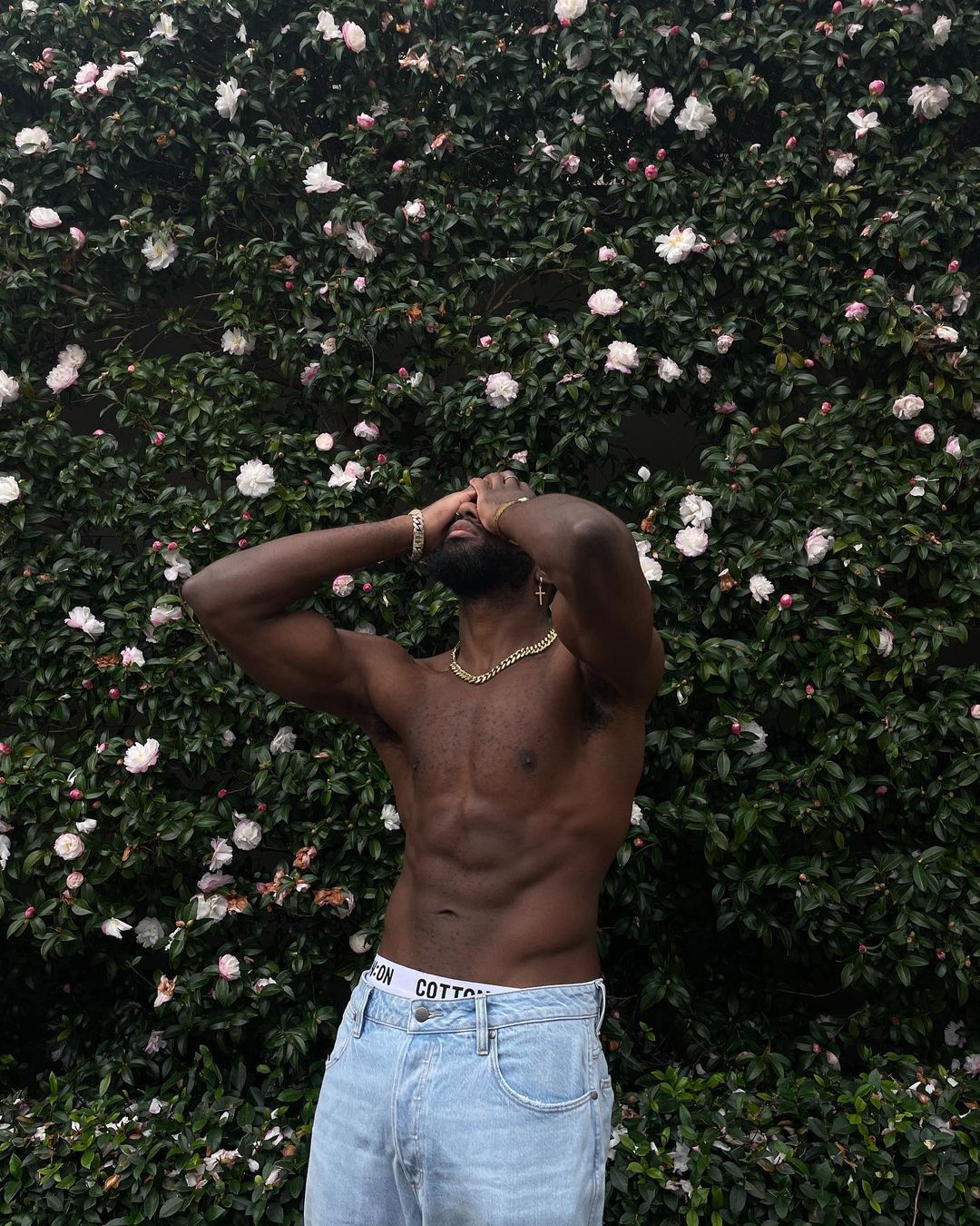Shirtless adult male poses in front of a flowering bush wearing jeans, a chain, and bracelets, with hands near his face.