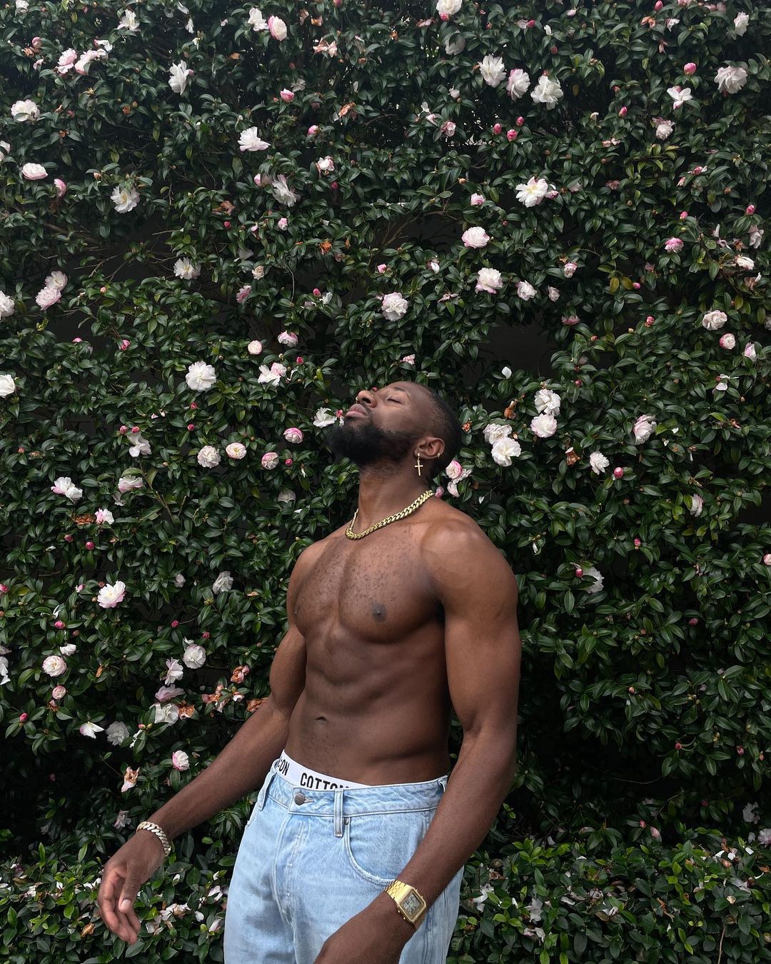 Shirtless Black man stands with closed eyes in front of a flowering bush, wearing jeans, gold chain, and a gold watch in daylight.