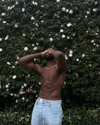 Shirtless black man wearing light wash denim jeans posing with hands on face against a dense green hedge of white flowers