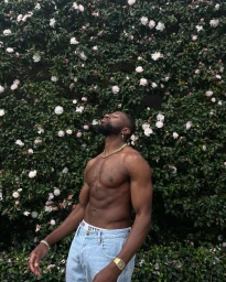 Shirtless black man wearing light wash denim jeans and gold jewellery standing before a dense flowering hedge