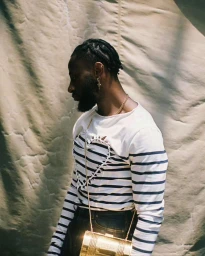 Black man in profile wearing a striped long-sleeve top and carrying a gold cylindrical crossbody bag against a textured backdrop