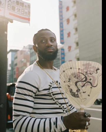 Black man with a beard wearing a striped long sleeve shirt holding a patterned Japanese folding fan on an urban street.