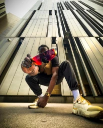 Low-angle shot of a black man squatting and holding red tinted shield sunglasses toward the camera in front of a tall modern building facade