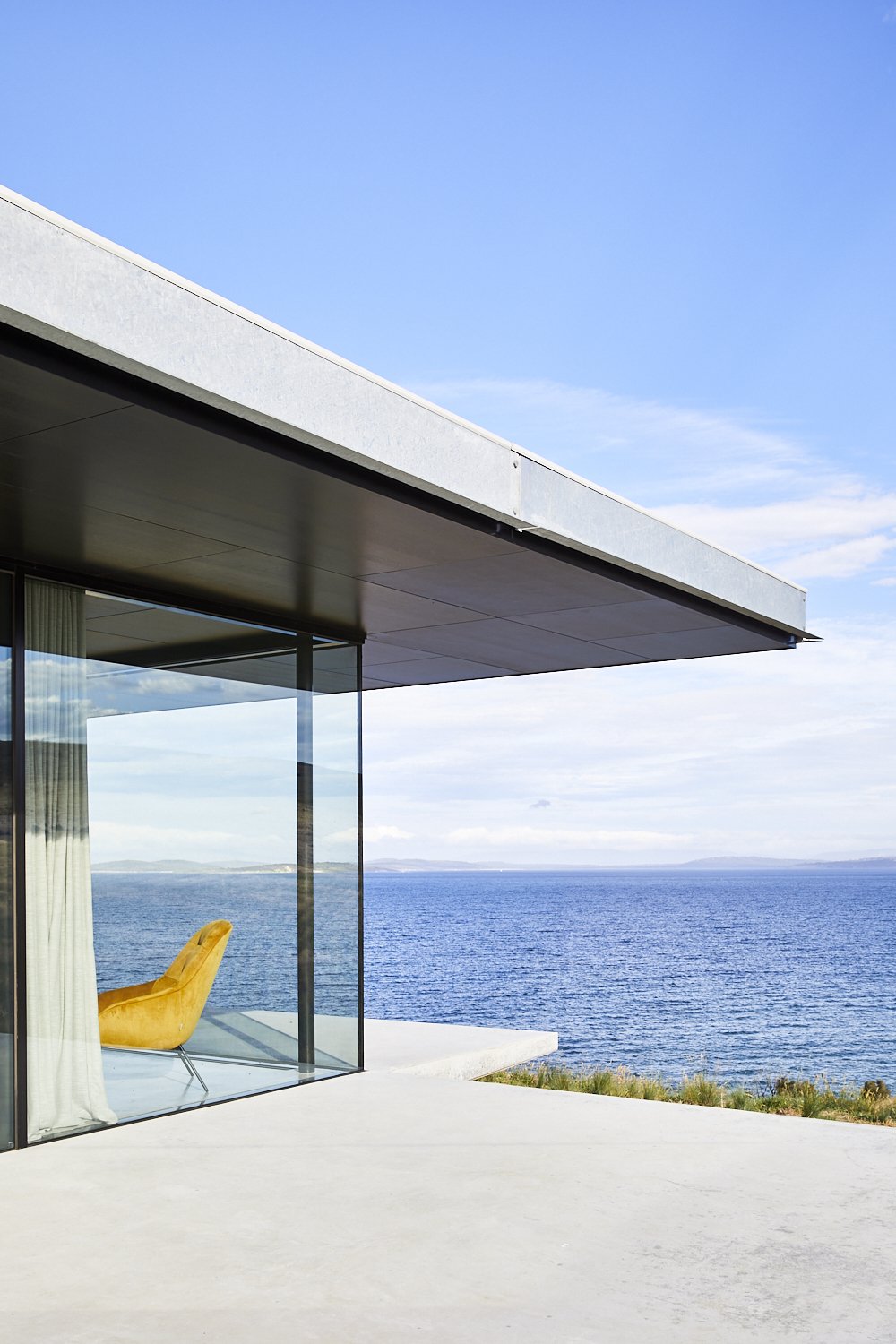 Yellow chair faces the ocean from the corner of a modern building with large windows and a concrete patio under a blue sky.
