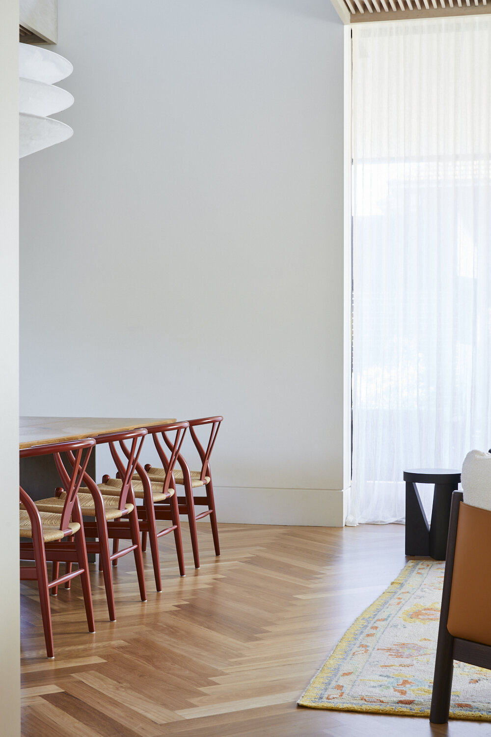 Minimalist dining room featuring red Wishbone chairs, light wood floor, white walls, modern chandelier and light filtering window.