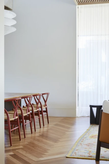 Minimalist dining room featuring red Wishbone chairs, light wood floor, white walls, modern chandelier and light filtering window.