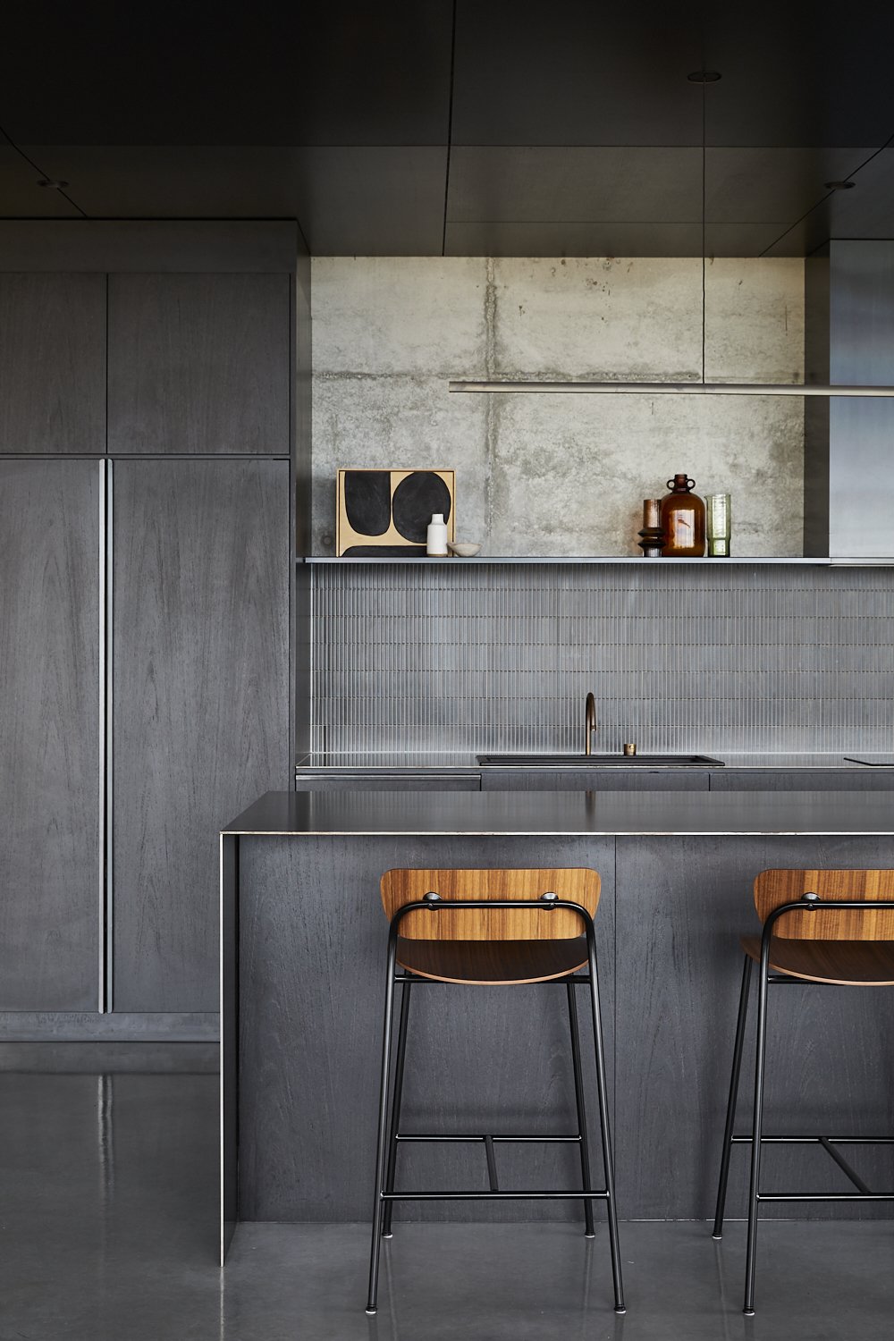 Minimalist kitchen design with grey cabinets, concrete walls, wood and metal bar stools, and natural light for a modern aesthetic.