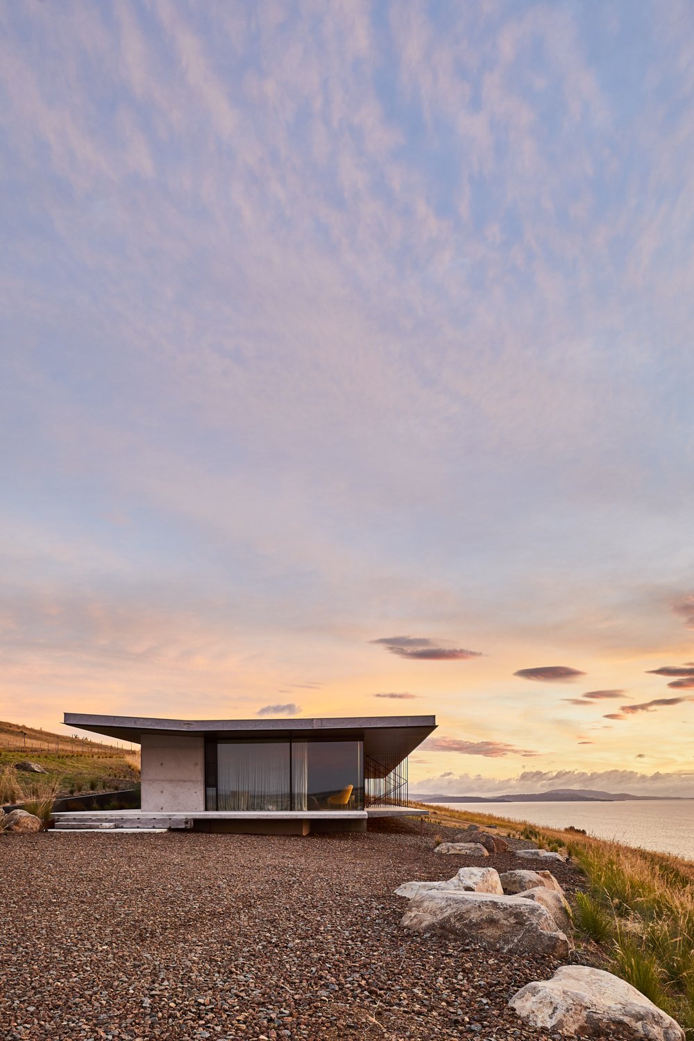 Modern architectural home with flat roof and glass wall, situated on a grassy hill overlooking the ocean at sunset with soft light.