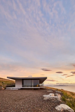 Modern architectural home with flat roof and glass wall, situated on a grassy hill overlooking the ocean at sunset with soft light.