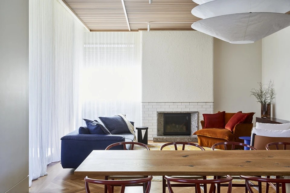 Living room interior featuring a fireplace, blue sofa, orange chair, and a wooden dining table with red chairs, bathed in natural light.