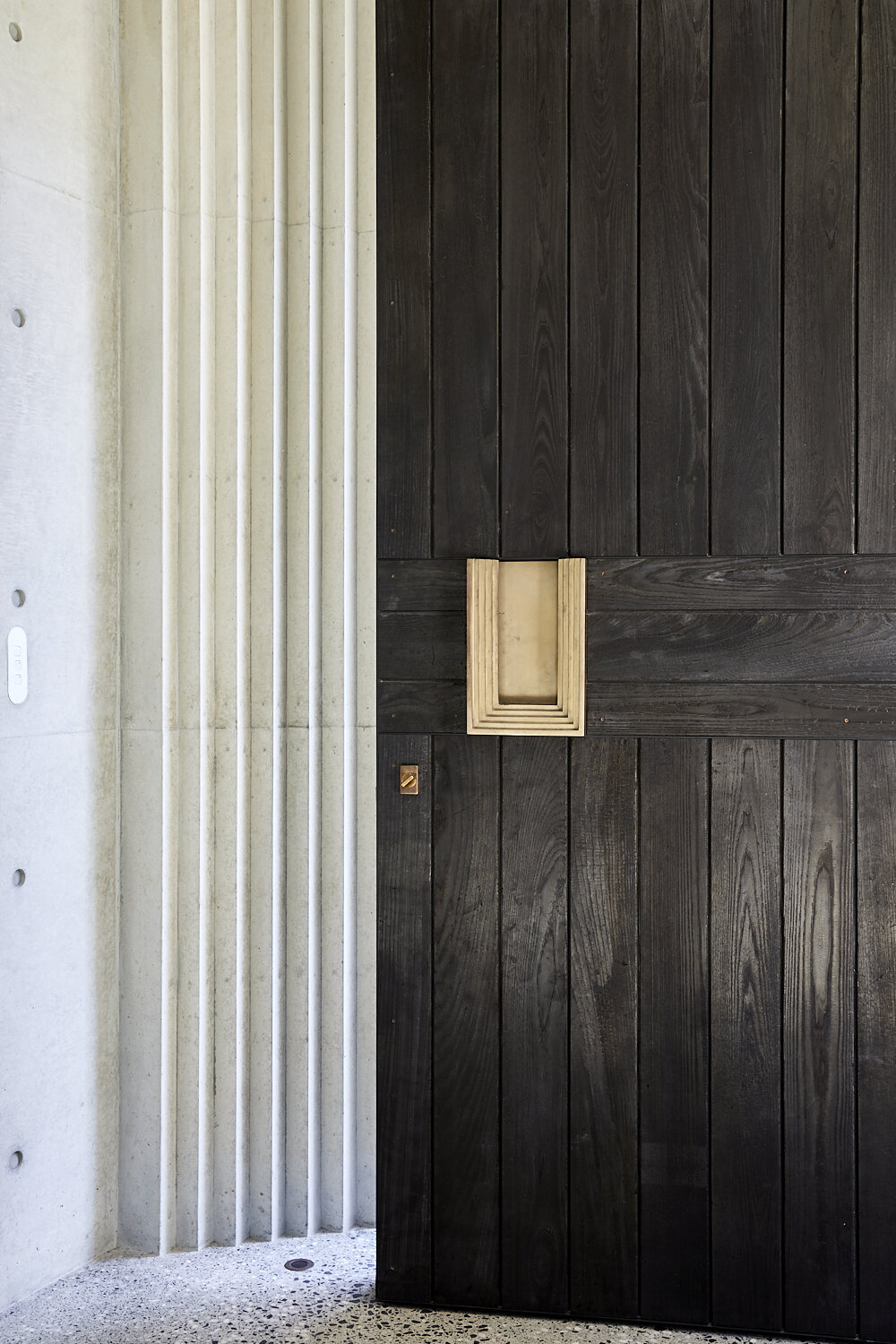 Architectural interior of an entryway with dark wood door with gold-colored hardware and a textured, light-colored concrete wall.