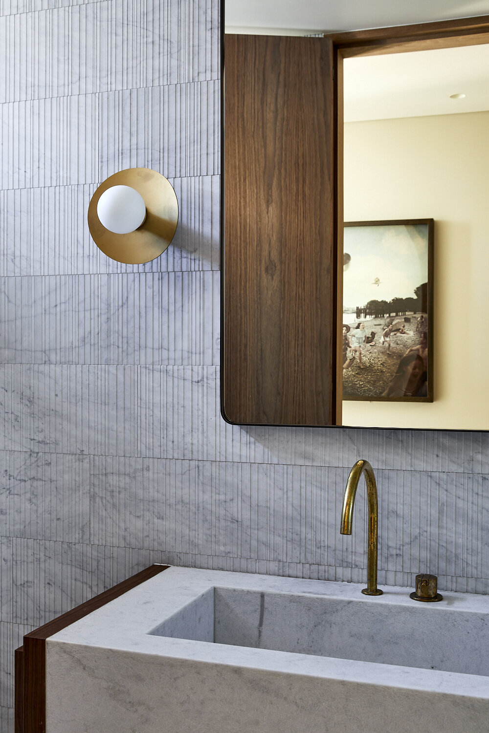 Modern bathroom interior with marble walls, brass faucet, and wood-framed mirror reflecting a framed artwork in a neutral setting.