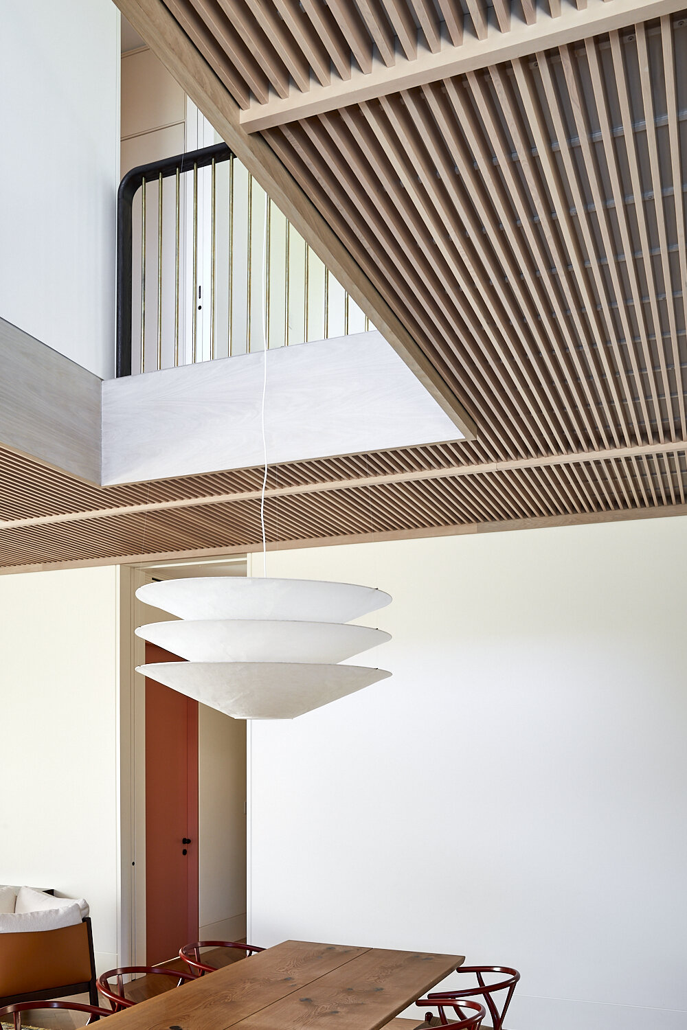 Interior shot showcases a modern pendant lamp, wooden ceiling slats, a wooden dining table, and red chairs in a minimalist space.